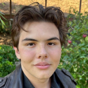 Selfie of a teenage boy with brown hair wearing a leather jacket in front of green bushes.