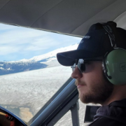 In a navy cap, black sunglasses and green over-the-ear headset, Hunter sits in the cockpit of a plane flying over clouds and looking out over the horizon.