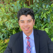 In a black suit, light purple shirt and red and blue tie, Bassam poses for a photo in front of a bright green bush.