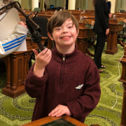 In a maroon-colored sweater, Dominic smiles for the camera as he holds a podium microphone at the California State Capitol.